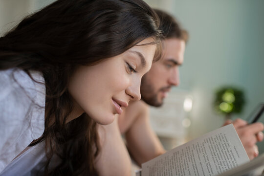 Woman Reading Book on the bed while her husband looking at his s