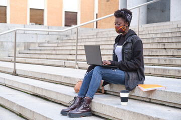 University female african student wearing protective face mask studying on her laptop sitting on stairs outside on campus. New normal in college.