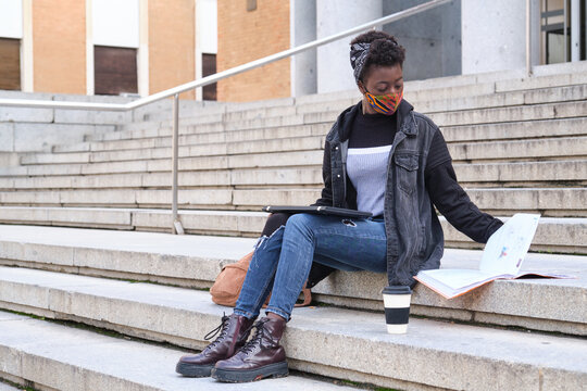 University female african student wearing protective face mask studying sitting on stairs outside on campus. New normal in college.