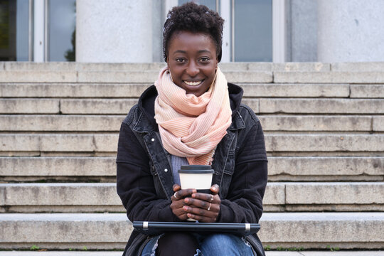 Beautiful university female african student smiling looking at camera, holding a cup of coffee, sitting on stairs outside on campus. College life concept.