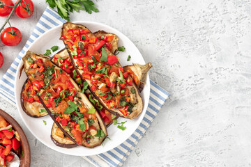 Baked eggplant with tomatoes, peppers, onions, parsley, and olive oil in a plate on a gray concrete table top view. Tasty vegetarian food Mediterranean style. Copy space for text