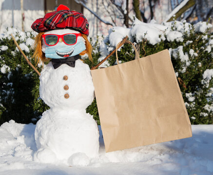 happy snowman in a bright red beret, sunglasses and a medical mask with a smile painted on it holds a large craft bag for the inscription of advertising text on it. Life during the COVID 19 pandemic