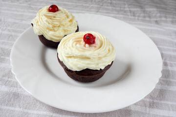 two gluten-free cupcakes with buttercream and red currant berries are placed in a white plate on a light side table . homemade cake