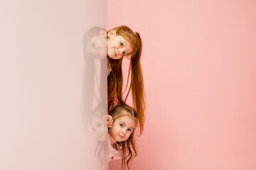 Greetings. Happy kids, girls isolated on coral pink studio background. Look happy, cheerful. Copyspace for ad. Childhood, education, emotions, facial expression concept. Peeking out from behind the
