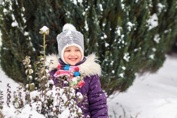 Warmly dressed little girl on a background of large thuja and snow. The child in the clothes looks away