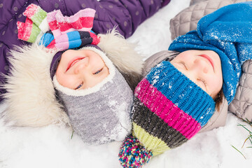 Two little girls are lying in the snow with their heads next to each other. Children lie in the snow, smiling and dreaming