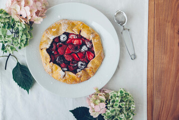 Delicious freshly baked vegan strawberry and cherry galette on wooden rustic background with hydrangea flowers, top view. Sweet food, summer dessert.