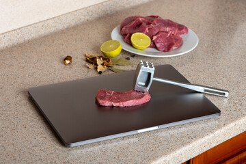 Computer is used as a kitchen board. Meat on laptop is ready to beat with tenderizer. Computer...