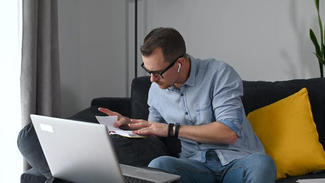 Focused and puzzled freelancer guy is doing paperwork remotely, analyst looks through documents working at home with laptop. An entrepreneur checks balance, expenses sitting on the couch