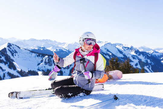 girl on skis sitting on snow. getting ready to take a selfy photo.