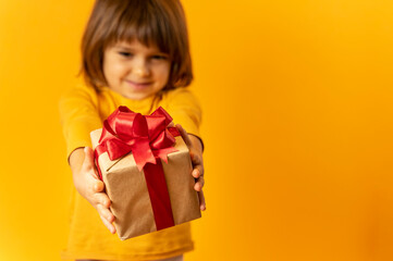 Cute Kid gives gift box with red ribbon on yellow background