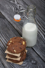 Oatmeal cookies with peanuts and chocolate. Folded in a column and laid out with paper. Nearby milk. On pine boards.