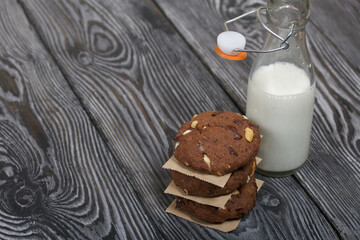 Oatmeal cookies with peanuts and chocolate. Folded in a column and laid out with paper. Nearby milk. On pine boards.