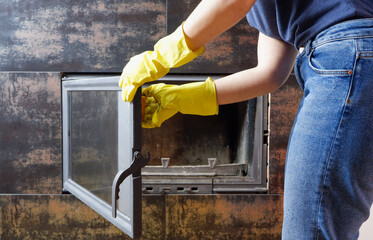 Close up of young woman in jeans, yellow gloves is cleaning the fireplace. Modern fireplace with glass.