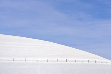 A white, snow-covered hill with footprints on it, with a fence, against a blue sky in winter