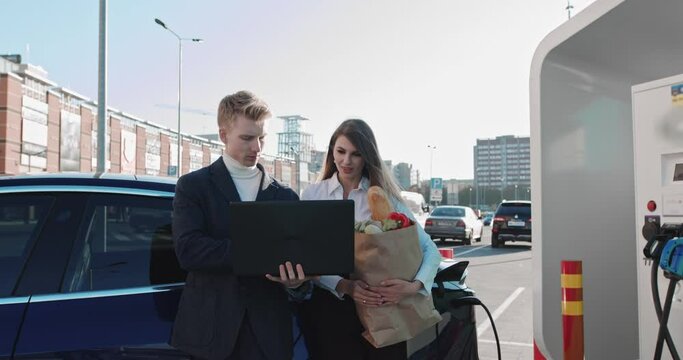 Man And Women After Shopping Products Work Near An Electric Car. Work remotely, Freelance. Car At The Charging Station. Successful People Work In The Yard. Work While Charging The Car.