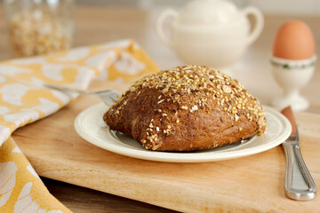 Closeup view of a healthy bun with multigrain mixed cereal seeds on a white porcelain plate with a knife, wooden tray and yellow dishcloth. Tasty breakfast with an boiled egg with selective focus.