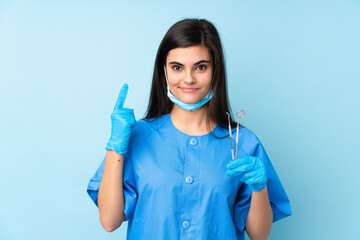 Young woman dentist holding tools over isolated blue background pointing with the index finger a great idea