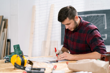 Portrait of a hardworking professional carpenter holding a angular ruler and pencil while measuring a board in a carpentry workshop. A bearded DIY enthusiast measures wood. There are a locksmith table