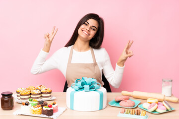Obraz na płótnie Canvas Pastry chef with a big cake in a table over isolated pink background showing victory sign with both hands