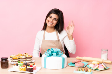 Obraz na płótnie Canvas Pastry chef with a big cake in a table over isolated pink background saluting with hand with happy expression