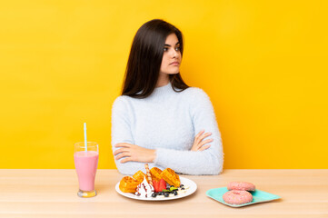Young woman eating waffles and milkshake in a table over isolated yellow background looking side