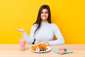 Young woman eating waffles and milkshake in a table over isolated yellow background holding copyspace imaginary on the palm
