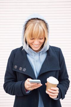 Happy blonde hair young woman in blue hoodie is using smartphone and drinking take away coffee while walking outdoors in the city. Happy face expression, smiling with teeth. 