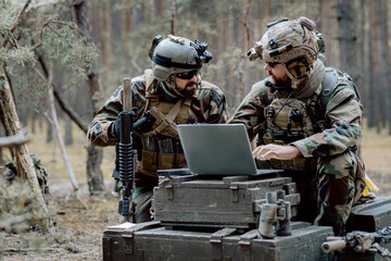 Bearded soldiers in uniform sit on military transport crates, analyze data on a laptop and work out...