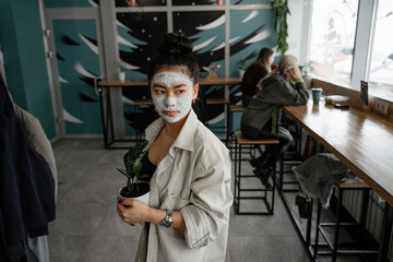 girl with a bun on her head and a clay mask on her face is holding a flower pot in a public place, in a cafe, the concept of feel at home