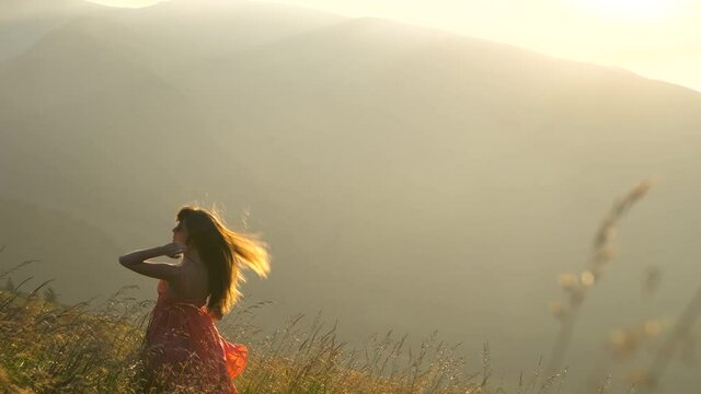 Young woman in red dress standing on grassy fild on a windy evening in autumn mountains enjoying view of nature.