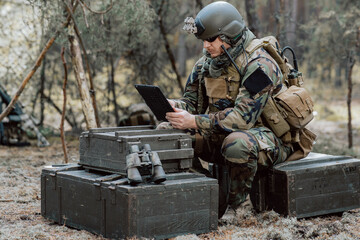 Bearded soldier in uniform sit on military transport crates, analyze data on a tablet and work out...