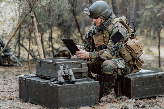 Bearded soldier in uniform sit on military transport crates, analyze data on a tablet and work out tactics at a temporary forest base. In the background, you can see a soldier protecting the base.