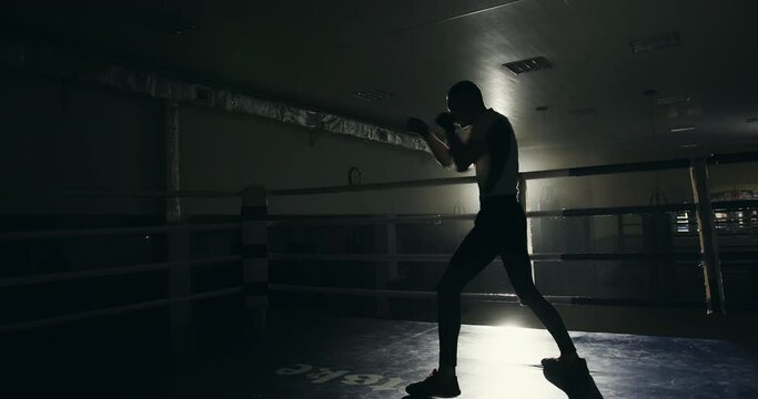 Professional male boxer training in the ring. Professional male boxer training in the ring in a darkened gym throwing punches and practicing his footwork backlit by a bright light