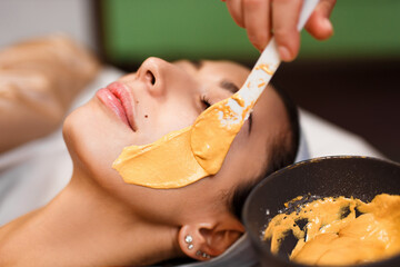 Procedure for skin care. Doctor in cosmetology office applies an yellow gold alginate mask to the face of young pretty woman.