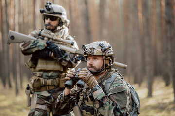 Portrait of a bearded middle-aged soldier in a Woodland military uniform and a helmet with headphones on his head, holding a rifle and looking around through the thick pine woods.