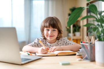 Distance learning online education. Caucasian smile kid boy studying at home with laptop, writing in notepad and doing school homework. Thinking child siting at table with notebook. Back to school.