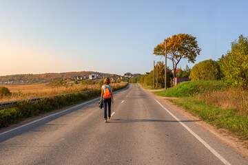 Fototapeta premium Back view of woman with backpack walking on autumn country road, hiking trip.