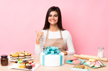 Obraz na płótnie Canvas Pastry chef with a big cake in a table over isolated pink background pointing to the side to present a product