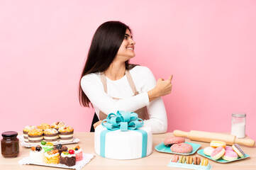 Pastry chef with a big cake in a table over isolated pink background pointing back