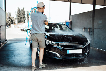Fototapeta premium Young man with hat washing his car during daylight at car wash station using high pressure water.