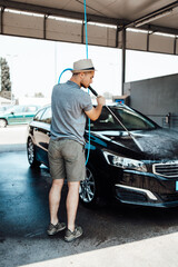Young man with hat washing his car during daylight at car wash station using high pressure water.