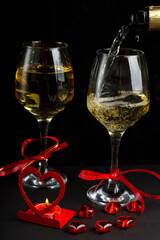 Champagne is poured into glasses on a black background next to a heart-shaped candlestick with a burning candle and a box with a ring.