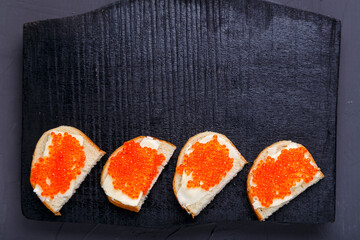 Bruschettes with butter and red caviar laid out on a black board on a gray concrete background copy space.