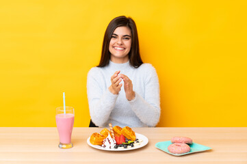 Young woman eating waffles and milkshake in a table over isolated yellow background applauding