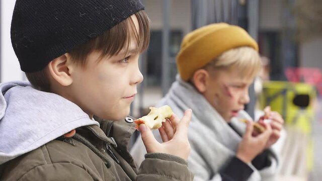 Homeless boys sit eating food on the bench, little caucasian children, street kids are hungry, get some food by kind people.