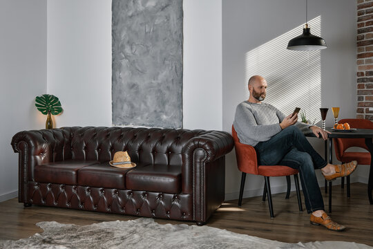 Portrait of an adult, bearded man in the interior of the living room, decorated in the loft style. Early morning, sunlight in the room. A mature man sits in a chair, reads messages on the phone.