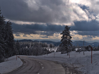 Stunning view of curvy pass road Schwarzwaldhochstraße in winter surrounded by deep snow and frozen trees near Schliffkopf peak, Germany in Black Forest with sun breaking through the cloudy sky.