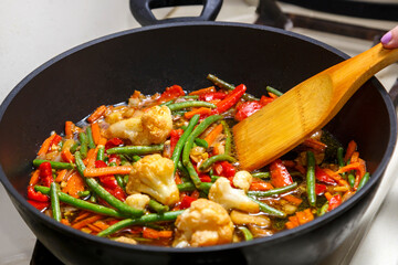 Vegetables in Pan-Asian sauce are stirred by a wooden spatula in a frying pan.
