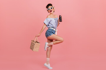 Full length view of dancing pinup girl. Studio shot of graceful young woman with bottle of beverage and bag.